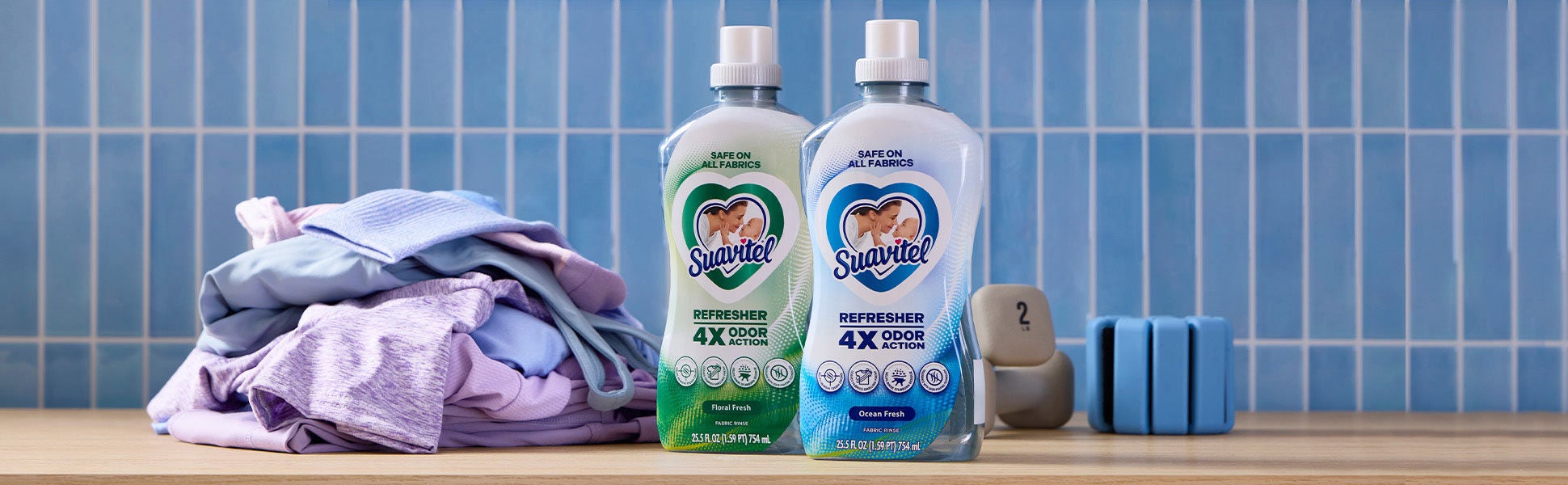 Dos botellas de Suavitel Refresher (aromas Floral Fresh y Ocean Fresh) sobre un mostrador de madera junto a una pesa de mano y ropa deportiva de color morado. Las etiquetas destacan '4X Acción Antiolor' y 'Seguro en todas las telas'.