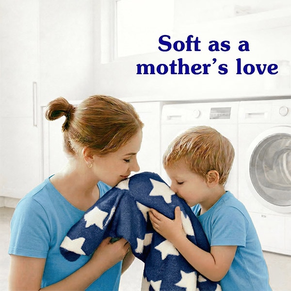 A mother and young son smiling and cuddling a soft navy blue blanket with white stars in a laundry room. Text reads: Soft as a mother's love.