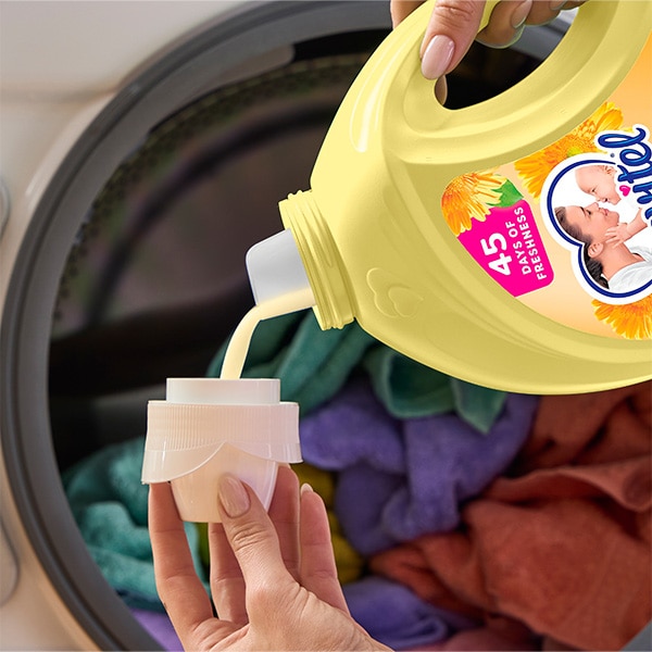 Pouring Suavitel Morning Sun fabric conditioner from yellow bottle into cap in front of a washing machine with colorful laundry Pouring Suavitel Morning Sun fabric conditioner from yellow bottle into cap in front of a washing machine with colorful laundry