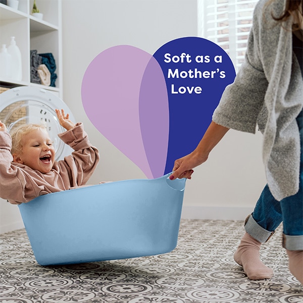 Smiling child sitting in a laundry basket as a mother pushes it, with heart-shaped graphic and textSoft as a Mother’s Love' in a laundry room. Smiling child sitting in a laundry basket as a mother pushes it, with heart-shaped graphic and textSoft as a Mother’s Love' in a laundry room.